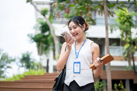 Asian woman office worker holding book talking on phone call while walking or standing in the park....
