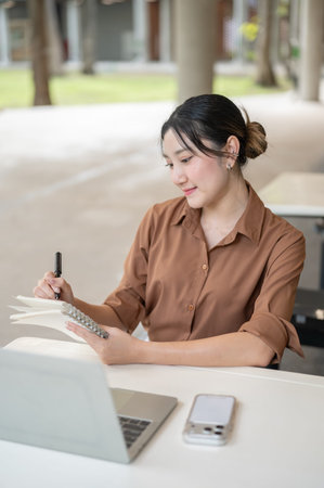 Asian woman student freelancer writing in notebook over laptop sitting at table in open air...