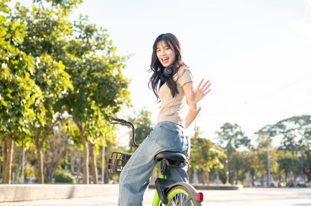 Happy asian woman waving hand while riding green rental bicycle on pavement in park or city square....