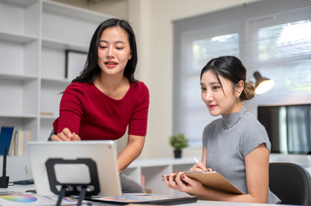 Pretty asian woman pointing on tablet and friend or coworker writing on clipboard at office's table. Interior Designer, Graphic Art, Modern Company.の写真素材