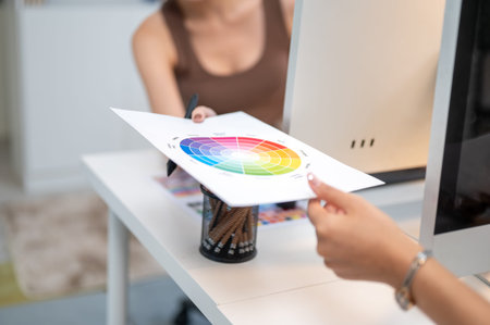 Close up of woman office worker's hand giving color chart to colleague over computer table in...