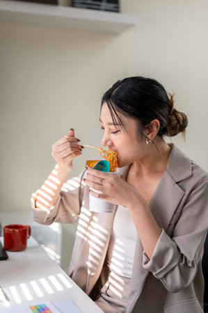 Hungry asian businesswoman or office worker eating instant noodle cup at table with bright sunlight....