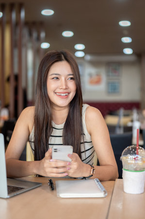 Pretty asian woman holding phone over notebook aside laptop and coffee while sits at table in cafe....