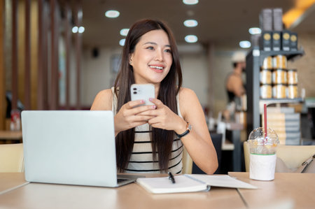 Pretty asian woman holding phone over laptop aside notebook and coffee while sits at table in cafe....
