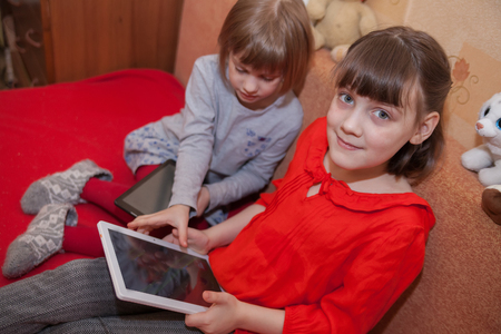 Two girls 5 and 7 years old playing tablets  at home on   couchの写真素材
