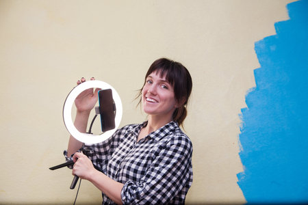 Woman in plaid shirt with ring lamp and smartphone on yellow background