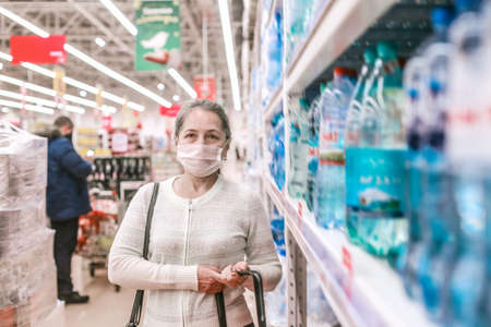 Senior female customer with shopping basket in supermarket wearing protective medical mask