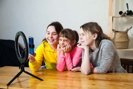 Three friends recording video on smartphone and lighting themselves with ring lamp at table in light roomの写真素材