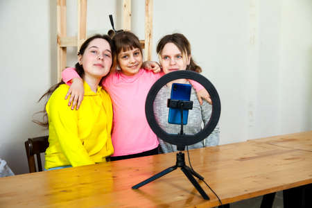 Three friends recording video on smartphone and lighting themselves with ring lamp at table in light roomの写真素材