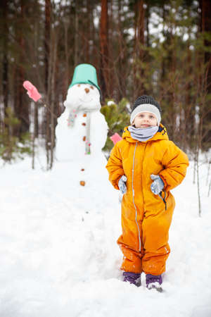 Girl in yellow jumpsuit in winter in forest near snowman.