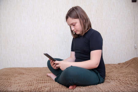 Portrait of pretty 12-year-old girl in black T-shirt and sweatpants on bed with tablet.の写真素材