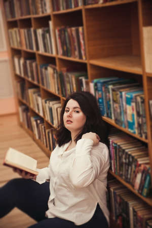 Young stylish woman reading book in library. Casual fashion, elegant look. Plus size model.の写真素材
