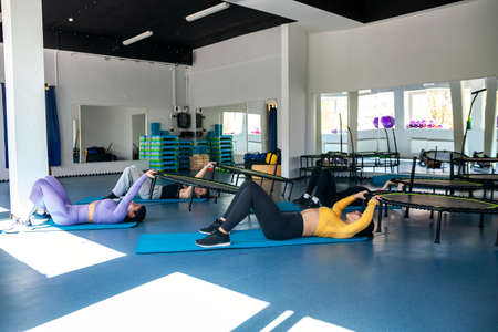 Three active women and fitness trainer in sport clothes doing exercises for jump with trampoline...