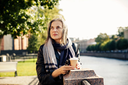 Well-groomed european elderly woman walking along promenade with paper cup of coffee in autumnの写真素材