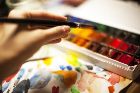 Artist illustrator's hand holds a brush with watercolor paints, on a white palette backgroundの写真素材