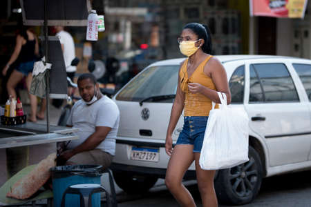 Rio de Janeiro, Brazil - July 3, 2020: People are wearing face masks during the Coronavirus pandemic in suburban area of the city. Woman with shopping bag passing while a man watches her.のeditorial素材