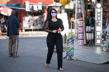 Rio de Janeiro, Brazil - July 3, 2020: People are wearing face masks during the Coronavirus pandemic in suburban area of the city. Woman dressed in black crossing the street.のeditorial素材