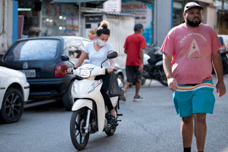 Rio de Janeiro, Brazil - July 3, 2020: Only some people are wearing face masks during the Coronavirus pandemic in suburban area of the city. Woman on a scooter in the street.のeditorial素材
