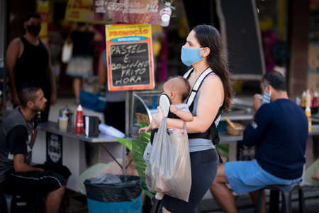 Rio de Janeiro, Brazil - July 3, 2020: People are wearing face masks during the Coronavirus pandemic in suburban area of the city. Mother carrying her baby and shopping bags.のeditorial素材