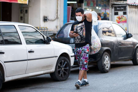 Rio de Janeiro, Brazil - July 3, 2020: People are wearing face masks during the Coronavirus pandemic in suburban area of the city. Man carrying big plastic bag on his back.のeditorial素材