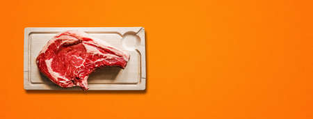 Raw beef prime rib and wooden cutting board isolated on orange background. Top view. Horizontal bannerの写真素材
