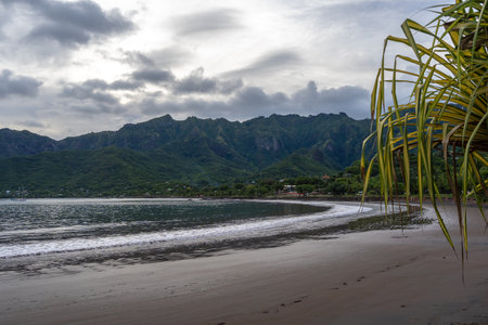 The black sand beach of Taiohae Bay in Nuku Hiva, Marquesas Islands, is framed by lush vegetation, coconut trees, and rugged green mountains under a cloudy sky. French Polynesiaの写真素材