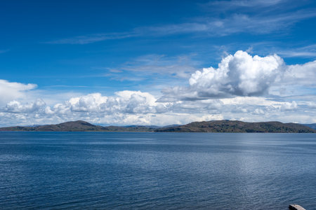 A peaceful view of Lake Titicaca from Llachon, Peru, with deep blue waters, scattered islands, and rolling hills under a bright sky with fluffy cloudsの写真素材
