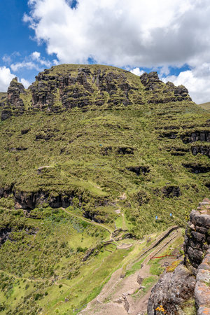 Green mountain ridges and steep rocky cliffs surround the Waqrapukara archaeological site, offering breathtaking panoramic views of the Andean landscape under a clear blue skyの写真素材