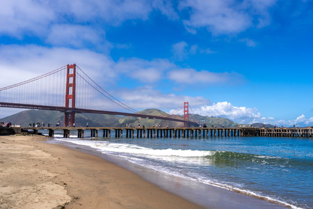 The Golden Gate Bridge stretches over the bay, viewed from the Beach. Waves lap against the shore...