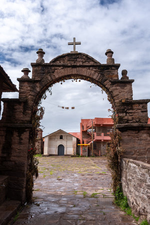 A stone arch with a cross and carved figures marks an entrance in a traditional village on Taquile Island, Lake Titicaca, Peru. The structure is decorated with dried flowers and local ornamentsの写真素材