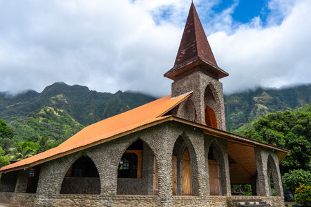 The Vaitahu catholic church in Tahuata, Marquesas Islands, French Polynesia, features a stone structure, wooden doors, and a tall steeple, set against lush green mountains under a cloudy skyの写真素材