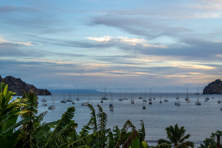 The sky over Taiohae Bay in Nuku Hiva, Marquesas Islands, glows with warm sunset colors, reflecting on the calm ocean, where sailboats are anchored near lush mountains. French Polynesiaの写真素材