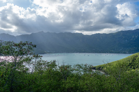 Taiohae Bay in Nuku Hiva, Marquesas Islands, is surrounded by green mountains under a blue and cloudy sky, with sailboats anchored in its calm waters near the coastal town. French Polynesiaの写真素材
