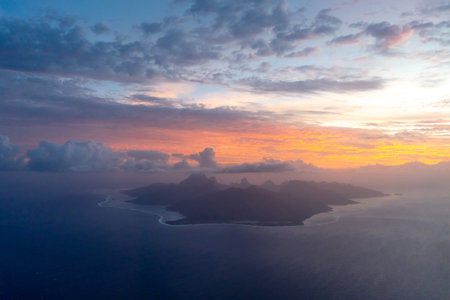 Moorea island, seen from above during sunset, reveals its rugged peaks, surrounding lagoon, and dramatic coastline, bathed in warm golden and blue huesの写真素材