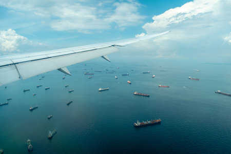 An airplane descends toward Singapore, flying above a dense gathering of cargo ships on calm blue...