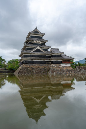 Matsumoto Castle stands majestically above its moat, surrounded by lush greenery and cloudy skies, embodying centuries of Japanese architectural eleganceの写真素材
