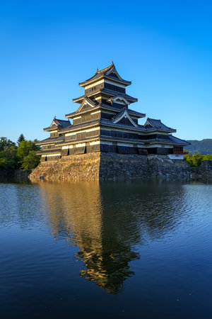 Matsumoto Castle stands majestically as golden light reflects off its dark wooden exterior and calm surrounding waters in Matsumoto, Japanの写真素材