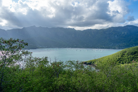 Taiohae Bay in Nuku Hiva, Marquesas Islands, is surrounded by green mountains under a blue and cloudy sky, with sailboats anchored in its calm waters near the coastal town. French Polynesiaの写真素材