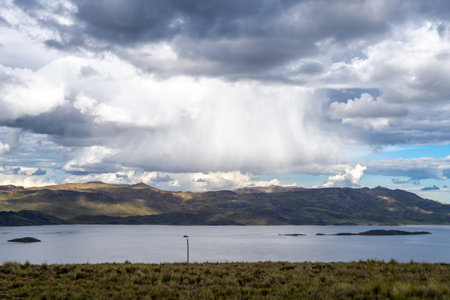 Lake Lagunillas in Peru is surrounded by rolling hills and grasslands, with dark storm clouds above and a distant rain shower falling over the mountainsの写真素材
