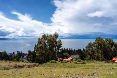 A small village on Amantani Island, Peru, with agricultural fields and scattered houses, overlooks the calm waters of Lake Titicaca under a sky filled with cloudsの写真素材