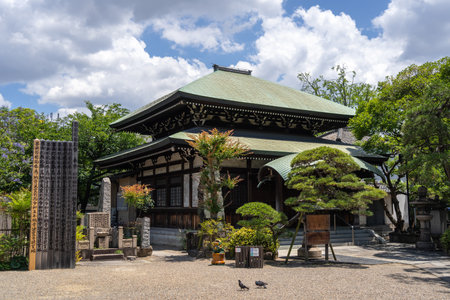The main hall of Isshin-ji Temple in Osaka is framed by sculpted trees and stone lanterns under a partly cloudy summer skyの写真素材
