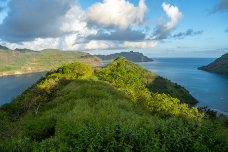 Golden light illuminates the lush green hills of Taipivai Bay in Nuku Hiva, French Polynesia, with the ocean stretching toward the horizon under a dramatic skyの写真素材