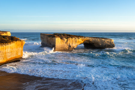 Golden light illuminates London Bridge, a famous rock formation at the Twelve Apostles site along the Great Ocean Road, Australia, with waves crashing against the cliffsの写真素材