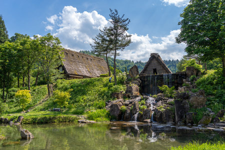 A small cascading waterfall flows into a peaceful pond surrounded by traditional gassho-style houses and lush greenery in Shirakawa-go, Japanの写真素材