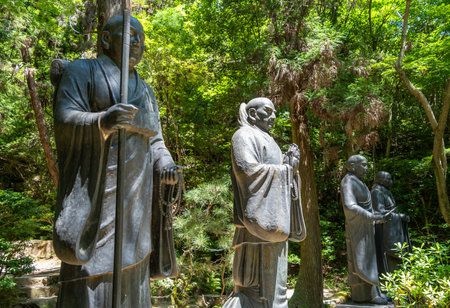 Tall bronze statues of monks stand solemnly in the forested grounds of Mitaki-Dera temple in Hiroshima, radiating peace and reverenceの写真素材