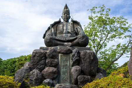 Bronze statue of shogun Minamoto no Yoritomo seated in armor on a stone base in Genjiyama Park, Kamakura, surrounded by greenery and skyの写真素材