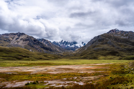 A marshy lake reflecting the dramatic cloudy sky is surrounded by rolling hills and snow-capped peaks on the Pastoruri Glacier trek in Huaraz, Peru. This high-altitude landscape offers breathtaking scenery, showcasing untouched Andean nature and glacial beautyの写真素材