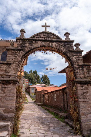 A stone arch with a cross and carved figures marks an entrance in a traditional village on Taquile Island, Lake Titicaca, Peru. The structure is decorated with dried flowers and local ornamentsの写真素材