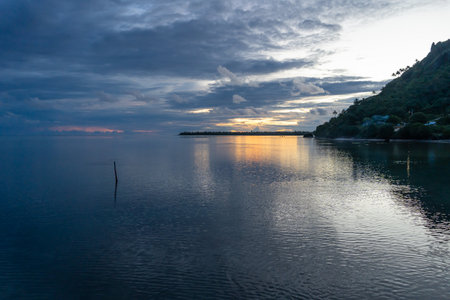 A golden sunset reflects on the calm waters of the Maupiti lagoon in French Polynesia, with dramatic...