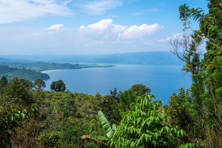 Wide view of Lake Poso surrounded by lush green hills and forests under a bright blue sky near...